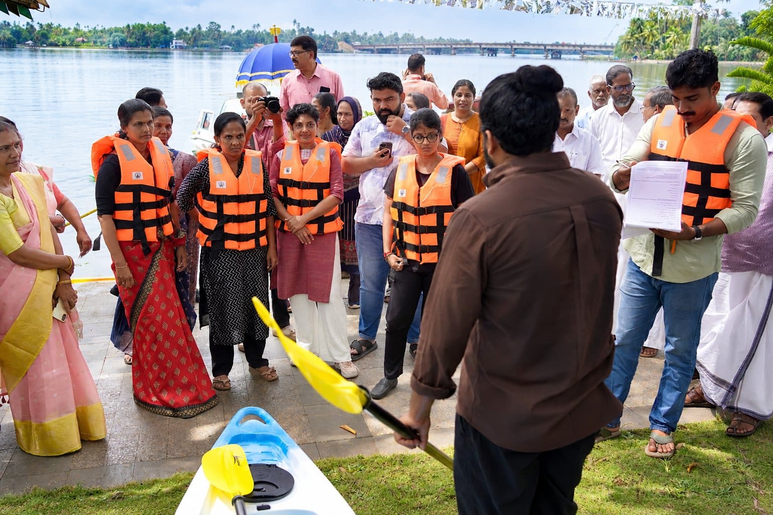 Guests in orange life jackets listening as a team member holds a paddle beside a kayak on the bank, with the backwater and bridge behind