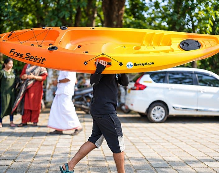 A team member carrying an orange kayak overhead across a paved waterside area
