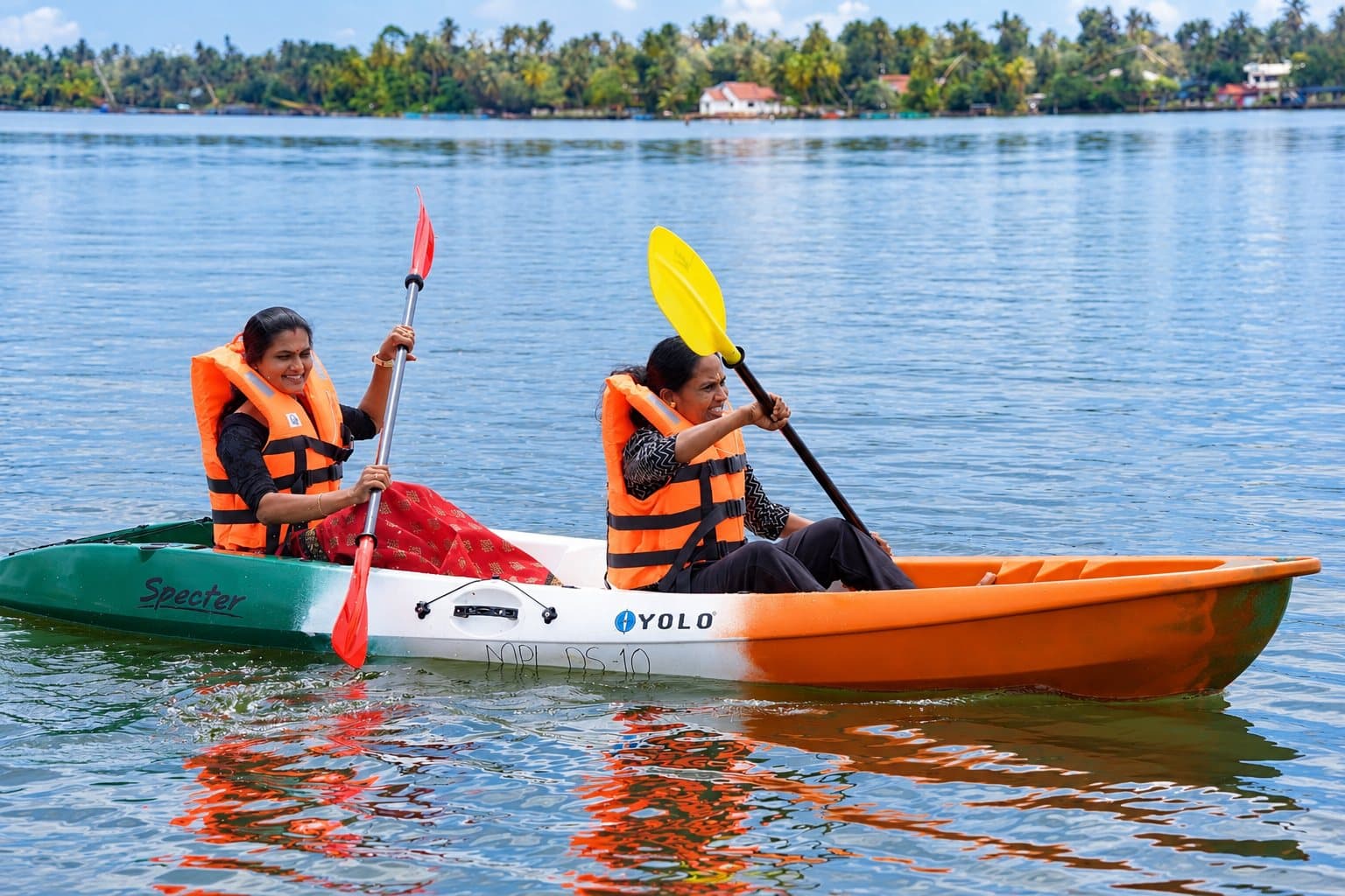 Two smiling women in orange life jackets paddling a green, white and orange tandem kayak on the backwater